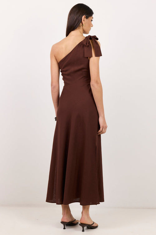 A woman modeling a dark brown one-shoulder midi dress with a bow detail on the shoulder, shown from the back against a plain white studio background.