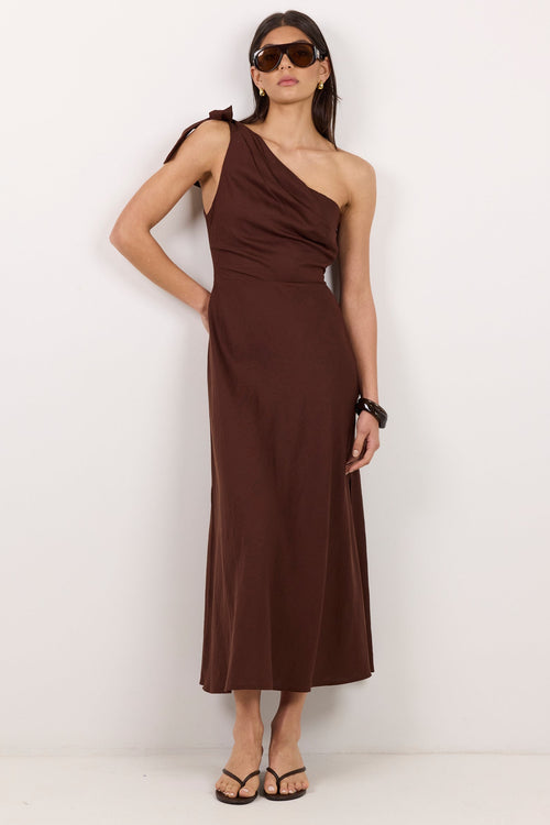 A woman modeling a dark brown one-shoulder midi dress with a draped neckline, standing against a plain white studio background.