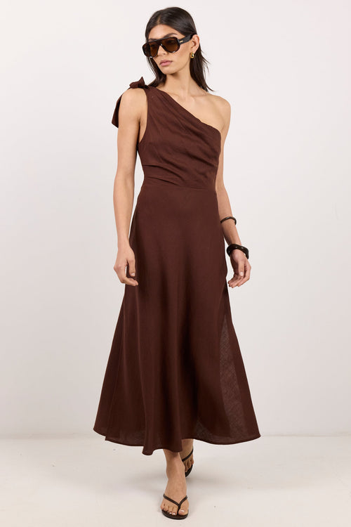 A woman modeling a dark brown, one-shoulder midi dress with a draped, gathered bodice and a flowing skirt, styled with sunglasses and flat sandals against a plain white studio background.