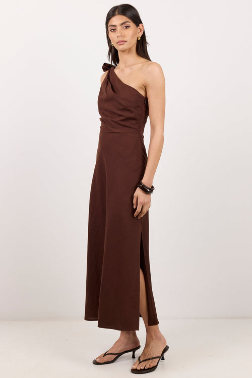 A woman modeling a dark brown, one-shoulder midi dress with a side slit, paired with black flat sandals, against a plain white studio background.