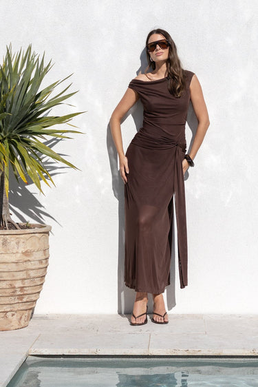A woman wearing a sleeveless brown maxi dress and sunglasses stands next to a white wall beside a pool, with a potted plant and trees in the background.