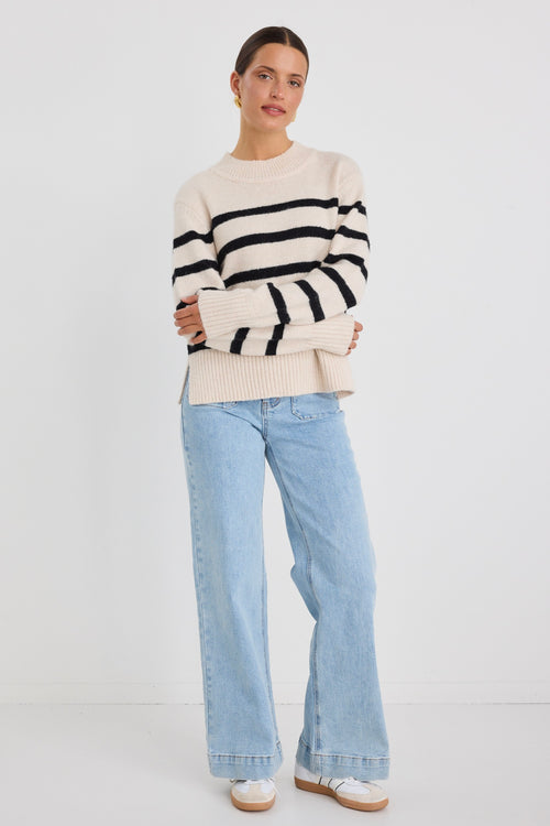 Woman wearing a cream and black striped funnel neck knit jumper, paired with light-wash wide-leg jeans and gold earrings.