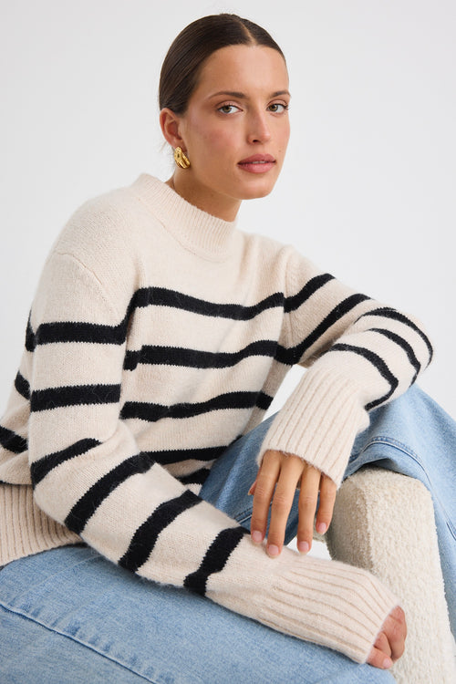 Woman sitting in a boucle chair wearing a cream and black striped funnel neck knit jumper, paired with light-wash wide-leg jeans and gold earrings.