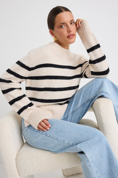 Woman wearing a cream and black striped funnel neck knit jumper, paired with light-wash wide-leg jeans and gold earrings.