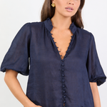 A woman modeling a navy blue short-sleeve blouse with a scalloped V-neckline and button front, paired with blue jeans against a plain white studio background.