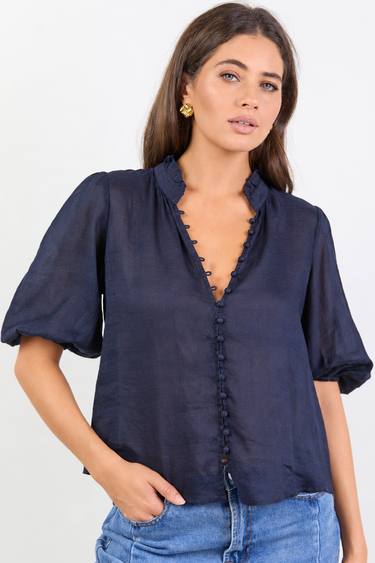 A woman modeling a navy blue short-sleeve blouse with a scalloped V-neckline and button front, paired with blue jeans against a plain white studio background.