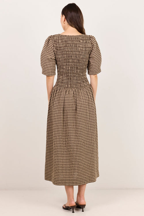A woman viewed from behind wearing a brown and beige houndstooth midi dress with puff sleeves and a smocked bodice, paired with black heels, against a plain off-white studio background.