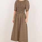A woman modeling a brown and beige checkered midi dress with puff sleeves and a smocked bodice, standing against a plain off-white studio background.