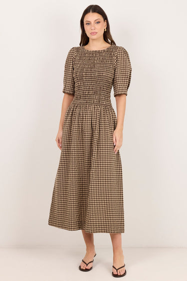 A woman modeling a brown and beige checkered midi dress with puff sleeves and a smocked bodice, standing against a plain off-white studio background.