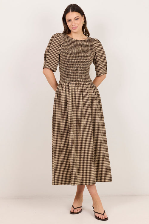 A woman modeling a brown and beige gingham midi dress with puff sleeves and a smocked bodice, standing against a plain off-white studio background.