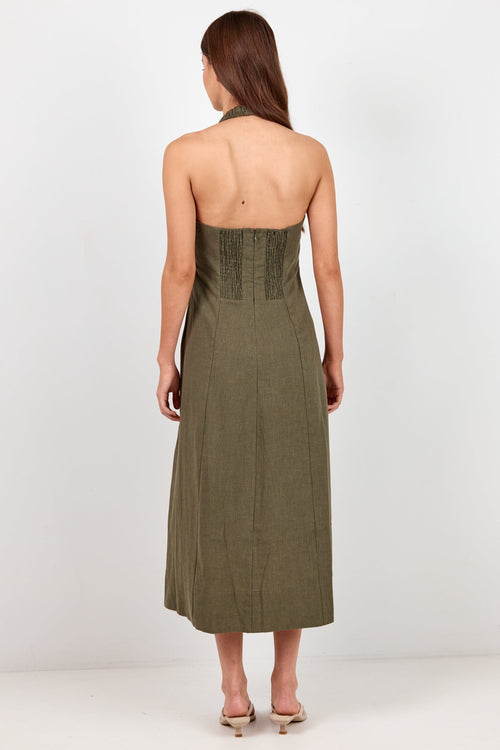 A woman viewed from the back wearing a sleeveless olive green halter midi dress with a structured corset-style waistband, standing against a plain white studio background.