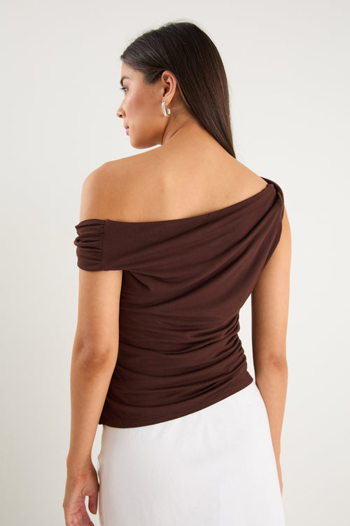 Woman wearing a chocolate brown off-shoulder sleeveless top with draped neckline, styled with white bottoms.