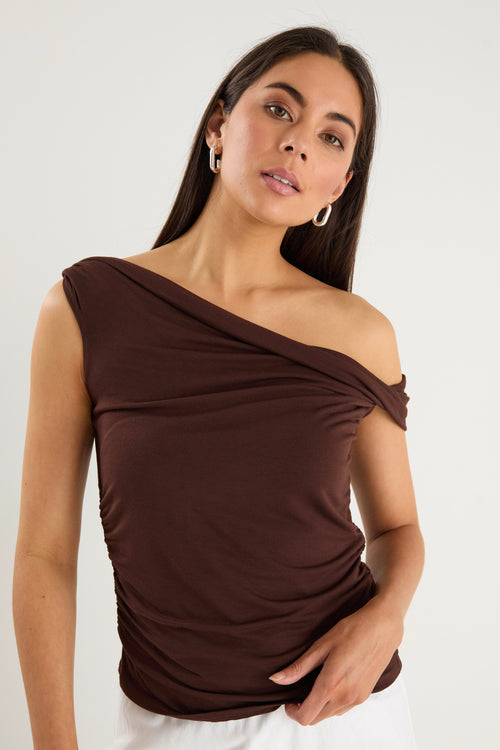 Woman wearing a chocolate brown off-shoulder sleeveless top with draped neckline, styled with white bottoms.