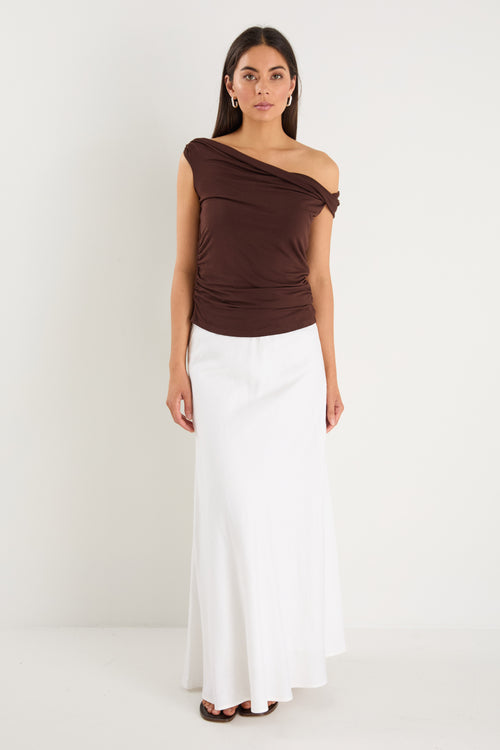 Woman wearing a chocolate brown off-shoulder sleeveless top with draped neckline, styled with white bottoms.