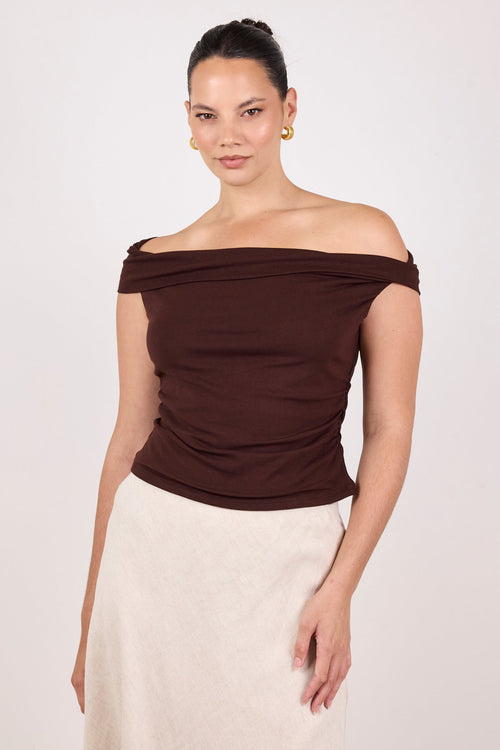 A woman modeling an off-the-shoulder ruched top in dark brown paired with a light beige skirt, against a plain light gray studio background.