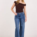 A woman modeling a dark brown off-the-shoulder top and blue wide-leg jeans against a plain white studio background.