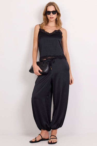 A woman modeling a black sleeveless camisole top with ruffled trim and matching black harem pants, styled with black sandals and sunglasses against a plain white studio background.
