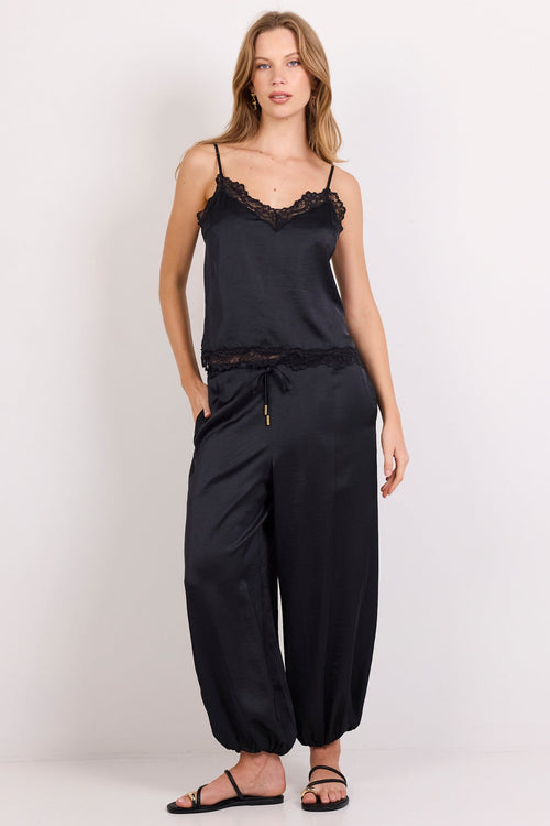 A woman modeling a black satin camisole top with ruffled lace trim and matching black wide-leg harem pants, styled with black strappy sandals against a plain white studio background.