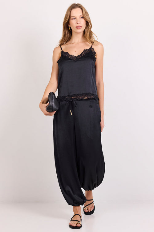 A woman modeling a black two-piece outfit consisting of a camisole top with lace trim and matching wide-leg harem pants, paired with black strappy sandals against a plain light gray studio background.