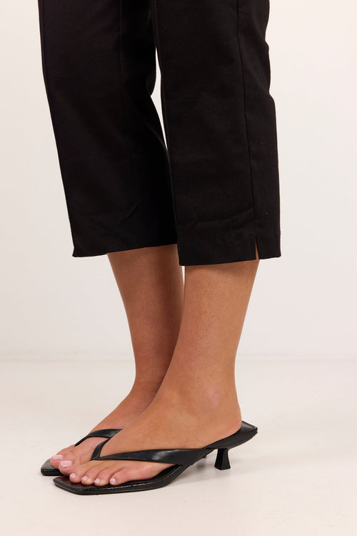 Black leather thong sandals with a low kitten heel, shown on a model wearing black cropped pants against a plain white studio background.