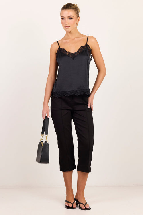 A woman modeling a black camisole top with lace trim, black cropped pants, and black sandals, holding a small black handbag against a plain white studio background.
