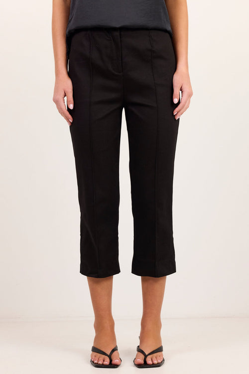 A model wearing black cropped trousers with a flat front and side pockets, paired with a dark gray top and black sandals, standing against a plain off-white studio background.