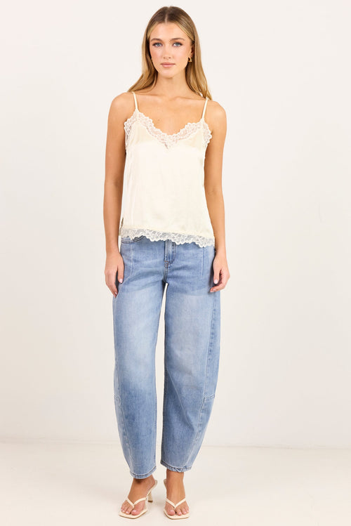 A woman modeling a cream lace-trimmed camisole top paired with light-wash wide-leg jeans and flat sandals, standing against a plain off-white studio background.