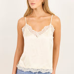 A woman modeling a cream-colored satin camisole top with delicate lace trim along the neckline and hem, paired with light-wash blue jeans against a plain off-white studio background.