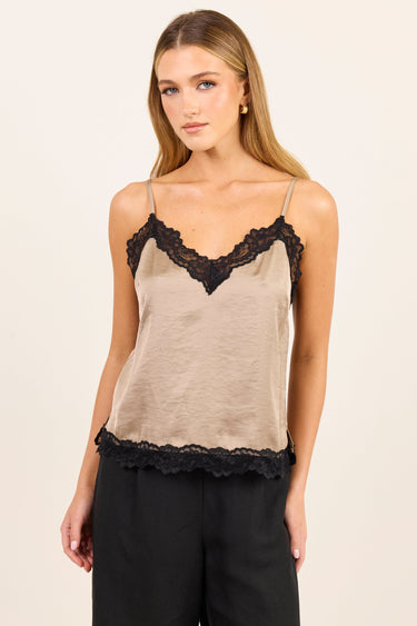 A woman modeling a beige camisole top with black lace trim along the neckline and hem, paired with black pants, against a plain light beige studio background.