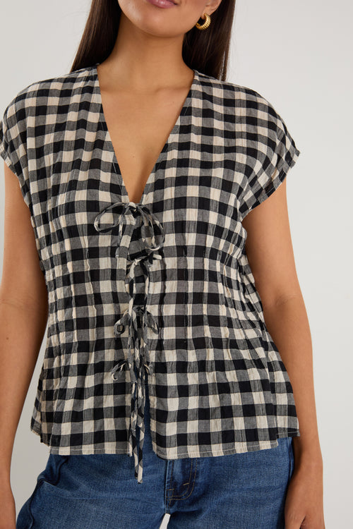 Woman wearing a sleeveless black and ecru gingham vest with tie-front detailing, styled with dark blue jeans and neutral flats.