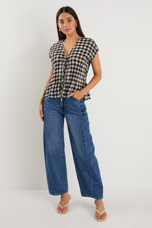 Woman wearing a sleeveless black and ecru gingham vest with tie-front detailing, styled with dark blue jeans and neutral flats.