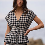 A woman modeling a matching black and white gingham check set consisting of a short-sleeve tie-front top and a long skirt, set against a blurred coastal background.
