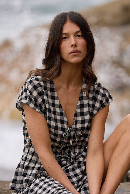 A woman sits on a large rock by the ocean, wearing a black and white gingham wrap dress with short sleeves and a tie at the neckline.
