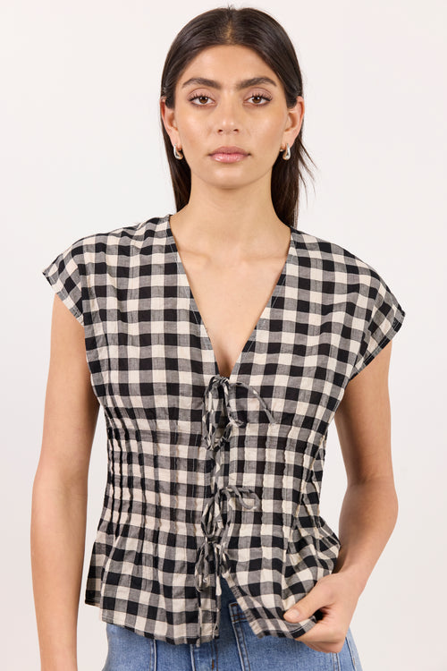 A woman modeling a black and beige gingham checkered top with a deep V-neck, tie-front detail, and cap sleeves, paired with blue jeans against a plain white background.