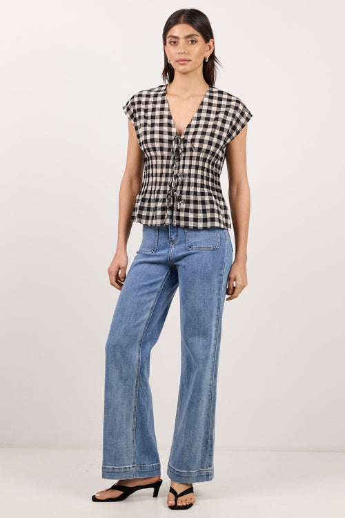 A woman modeling a black and white gingham check short-sleeve top with a front tie detail, paired with wide-leg blue jeans and black sandals, standing against a plain white studio background.
