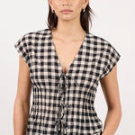 A woman modeling a black and white gingham checkered top with a deep V-neckline and front tie details, paired with light blue jeans, against a plain off-white studio background.