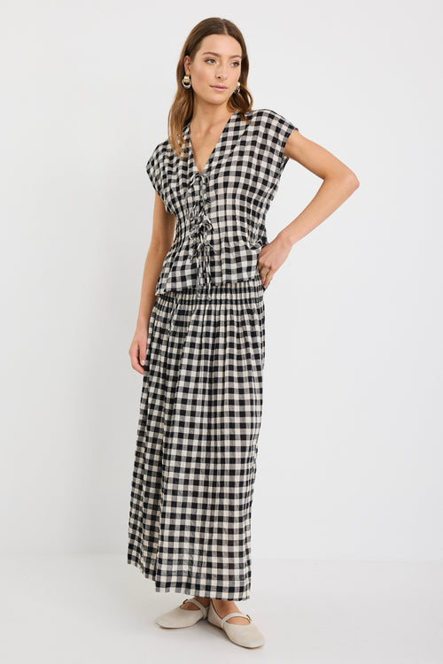 Woman wearing a sleeveless black and ecru gingham vest with tie-front detailing, styled with a matching long gingham skirt and neutral flats.