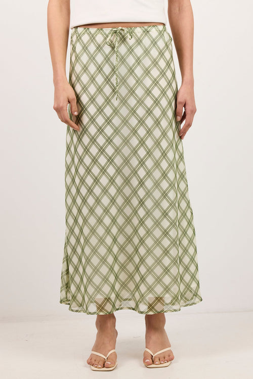 A woman models a long, flowing midi skirt with a green and beige plaid pattern, paired with a white top and white sandals against a plain white studio background.