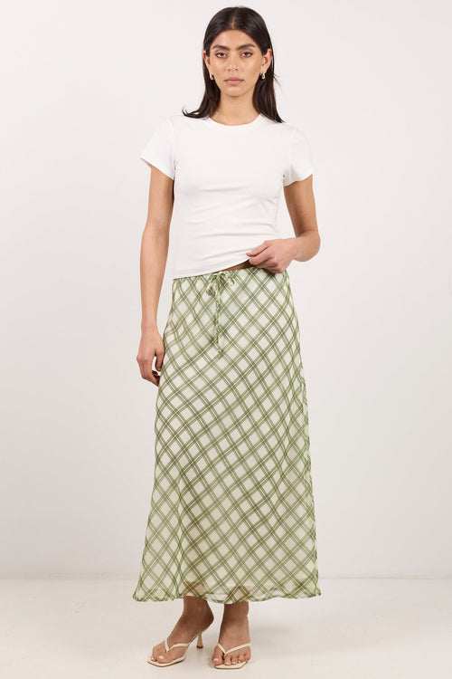 A woman modeling a long green and white gingham check maxi skirt with a drawstring waist, styled with a plain white t-shirt and beige platform sandals against a white studio background.