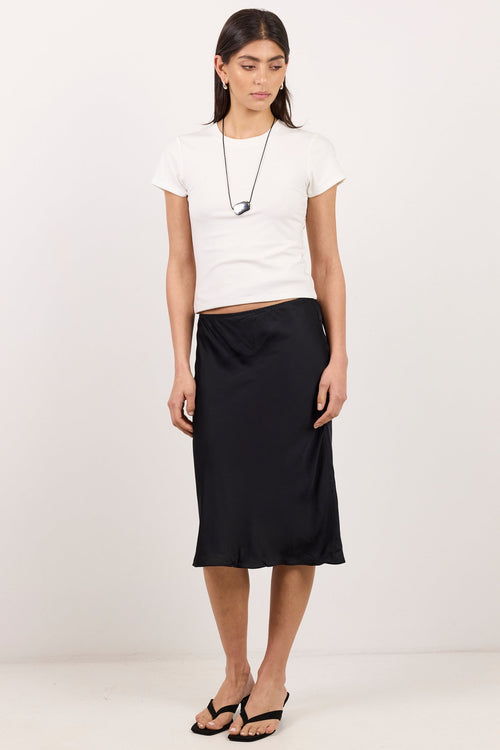 A model stands against a plain white studio background wearing a white cropped short-sleeve T-shirt, a black satin midi skirt, and black sandals.