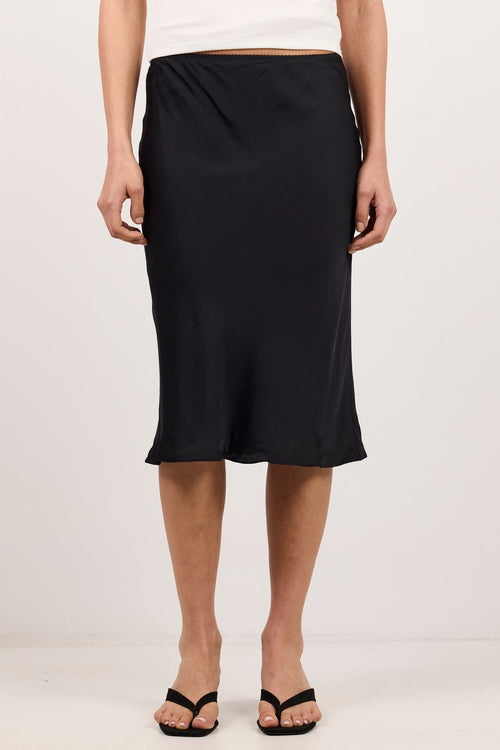 A model wearing a black midi skirt with a draped, gathered waist, styled with a white tank top and black sandals against a plain white studio background.