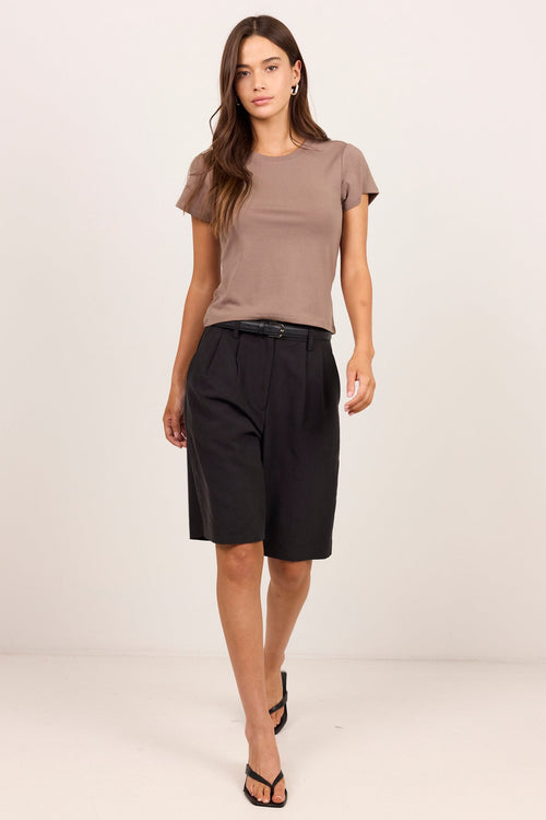 A woman modeling a taupe short-sleeve crewneck t-shirt and black knee-length pleated shorts with a black belt, styled with black flat sandals, against a plain off-white studio background.