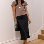 A woman stands indoors against a white paneled wall on a wood floor, wearing a taupe short-sleeve top, a black midi skirt, black sandals, a white patterned headscarf, sunglasses, and carrying a black handbag.
