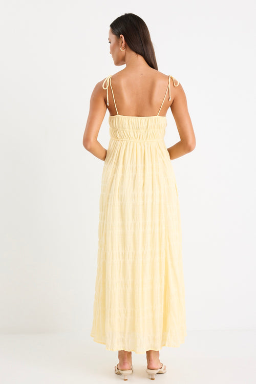 Woman wearing a pale yellow shirred maxi dress with thin tie-up straps and a flowing tiered skirt, paired with neutral sandals.