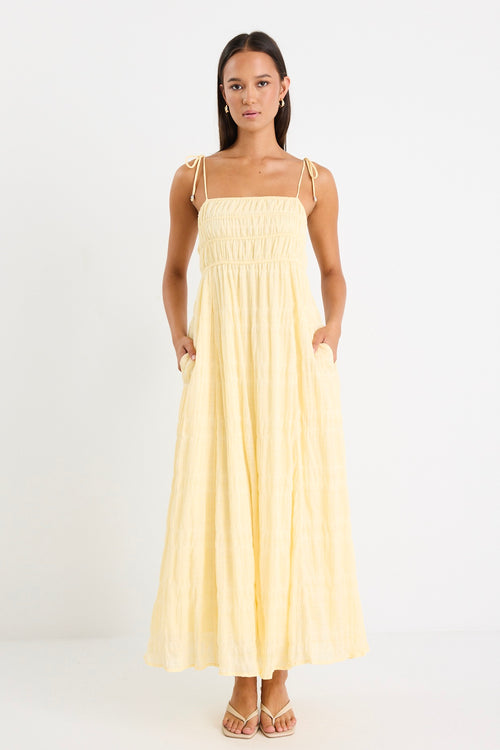 Woman wearing a pale yellow shirred maxi dress with thin tie-up straps and a flowing tiered skirt, paired with neutral sandals.