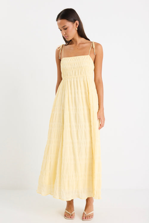 Woman wearing a pale yellow shirred maxi dress with thin tie-up straps and a flowing tiered skirt, paired with neutral sandals.