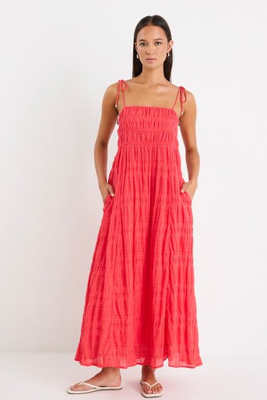 Woman wearing a poppy red shirred maxi dress with tiered detailing and tie-up straps, styled with white sandals.