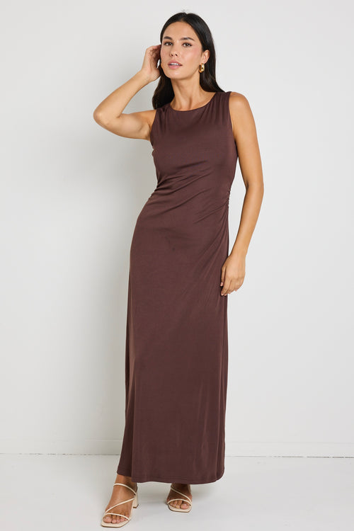 A woman wearing a chocolate brown sleeveless maxi dress with side ruching, styled with gold earrings and strappy nude heels.