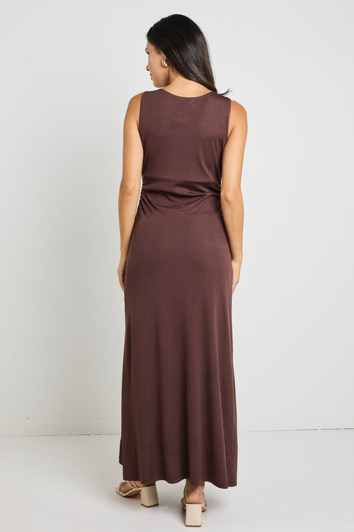 A woman wearing a chocolate brown sleeveless maxi dress with side ruching, styled with gold earrings and strappy nude heels.