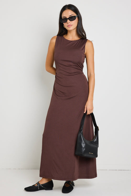 A woman wearing a chocolate brown sleeveless maxi dress with side ruching, styled with gold earrings and strappy nude heels.
